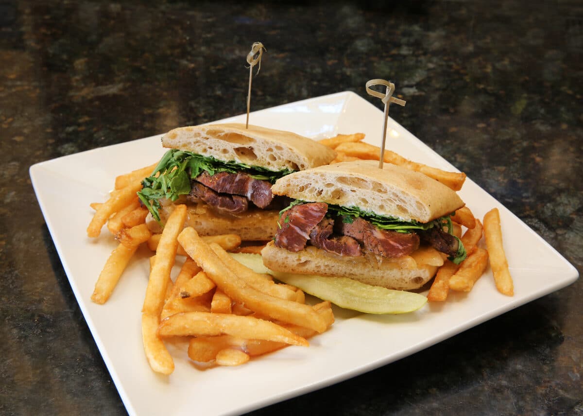 Steak Sandwich on ciabatta bread with flank steak, arugula, caramelized onions, and garlic horseradish sauce, served with fries.